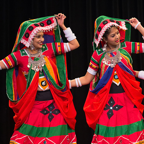 Two women dancers in colorful outfits, headdresses, and large jewelry leaning to the side.