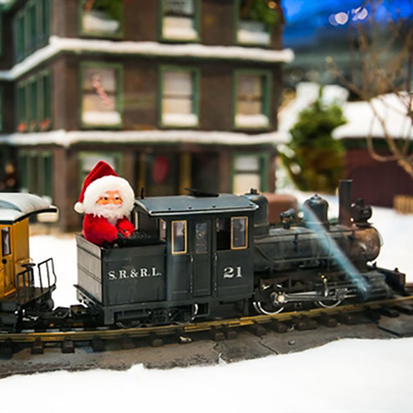 A Santa Clause figurine rides a model train car through an old fashioned village.